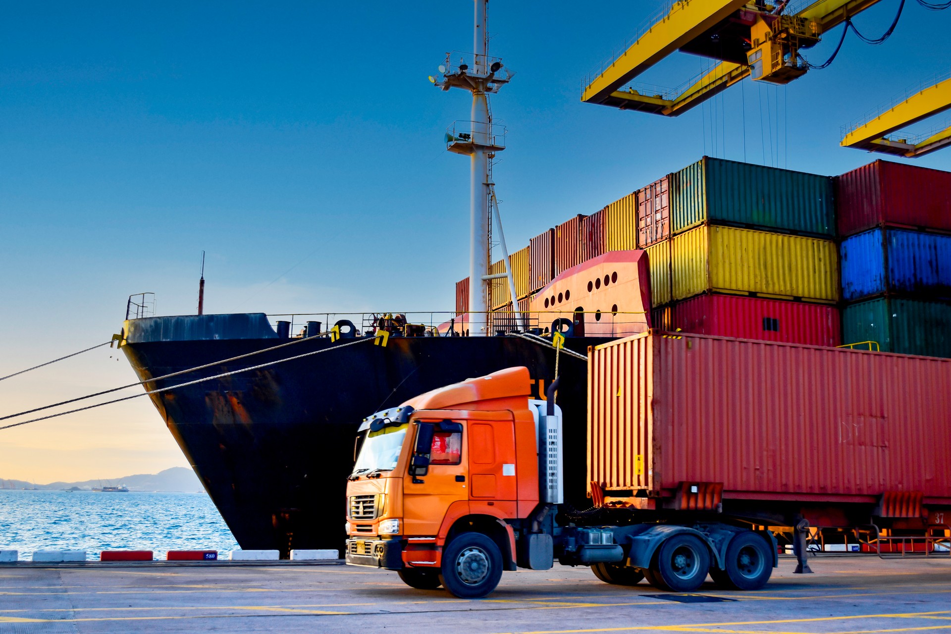 Truck carrying forty-foot container leaving port terminal with ship and quay crane on the background. Seaport operation activities, container shipping, and logistics. Truck carrying forty-foot container leaving port terminal with ship and quay crane on the background. Seaport operation activities, container shipping, and logistics.